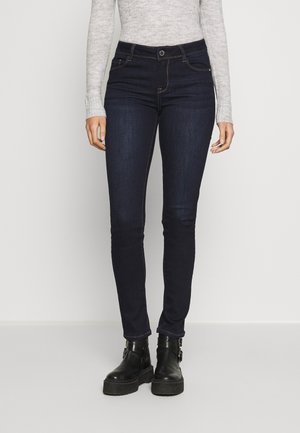 Jeans skinny de color azul oscuro con una textura suave, diseño de cinco bolsillos y costuras en contraste. Se combinan con botas negras de tobillo.
