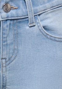 Primo piano di jeans di denim azzurro chiaro che mostrano la tasca frontale, il passante per cintura, le cuciture ricamate e il bottone in metallo.