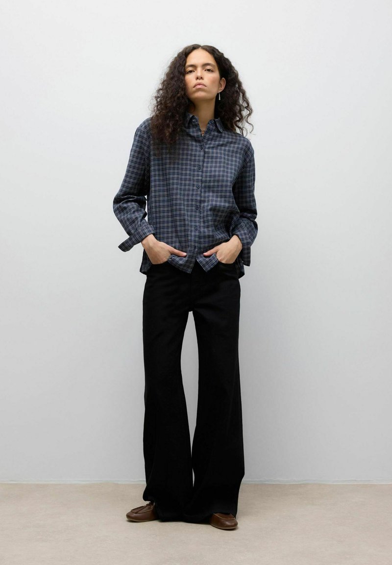 Een overhemd met knopen in marineblauw met een ruitpatroon, gecombineerd met zwart wijde broek en bruine loafers, tegen een neutrale achtergrond.