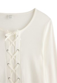 Lindex hvid ribbet top med rund halsudskæring og snøredetalje foran med metaløjer. Blød tekstur, lange ærmer.