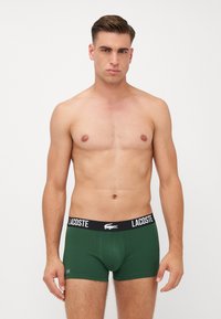 Boxers verdes con una banda negra que presenta la marca "Lacoste" en blanco. Tejido suave y liso, diseño ajustado sin patrones.