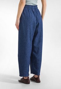 Osoba nosząca luźne, granatowe spodnie z wysokim stanem i elastycznym pasem oraz brązowe buty na sznurowadła, stojąca na białym tle.