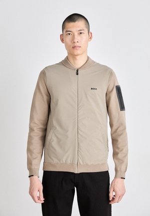 Chaqueta beige de cremallera con un frente liso, mangas acanaladas y un bolsillo decorativo. Presenta un cuello redondo y un logo en el pecho.
