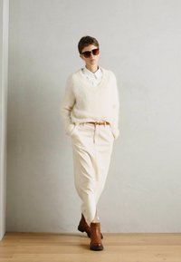 Pull en maille floue crème avec décolleté en V, associé à un pantalon blanc et des bottines brunes. Modèle portant des lunettes de soleil, se tenant devant un mur clair.