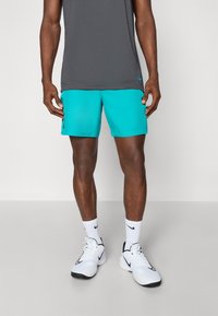 Shorts de sport turquoise et haut sans manches gris, portés avec des baskets blanches et des chaussettes. Les shorts présentent un logo noir sur le côté.