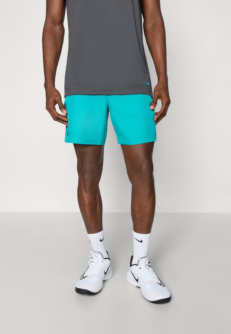 Shorts de sport turquoise et haut sans manches gris, portés avec des baskets blanches et des chaussettes. Les shorts présentent un logo noir sur le côté.