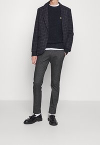 Veste à carreaux bleu marine sur un pull texturé sombre, pantalons gris sur mesure et chaussures en cuir verni noir à bout arrondi.