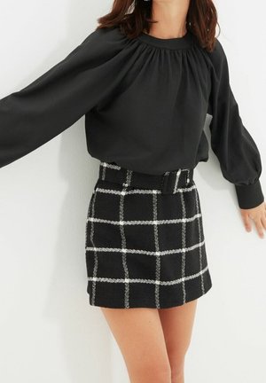 Vrouw die een zwarte blouse met lange mouwen en een zwart-witte geruite rok met hoge taille en riem draagt, poserend tegen een witte muur.