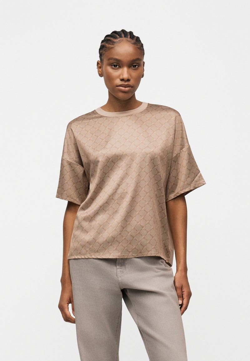 Γυναίκα με πλεκτά μαλλιά που φοράει μπεζ πουκάμισο από σατέν με μοτίβο και κοντά μανίκια και ανοιχτόχρωμο γκρι παντελόνι, στέκεται μπροστά σε λευκό απλό φόντο.