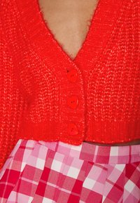 Roter gestrickter Cardigan mit tiefem V-Ausschnitt und vier herzförmigen Knöpfen. Darunter ist ein rosa karierter Rock mit helleren Karomustern sichtbar.