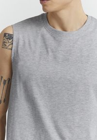 Graues ärmelloses Shirt aus einem weichen, strukturierten Stoff; mit einem gerippten Ausschnitt. Teilansicht eines Arms mit einem Tattoo.