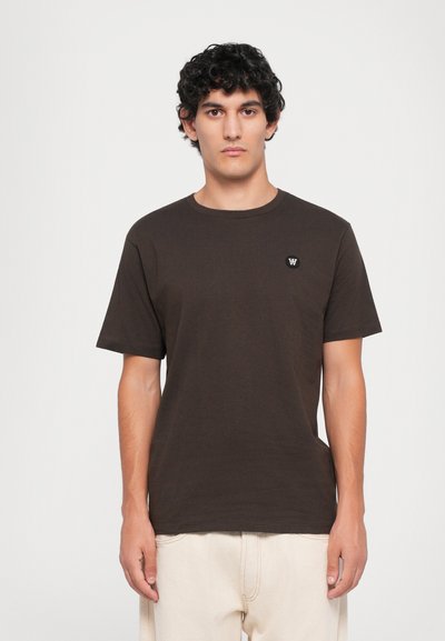 Homme avec des cheveux bouclés et foncés portant un T-shirt à manches courtes marron foncé uni avec un petit logo blanc sur la poitrine et un pantalon beige clair.