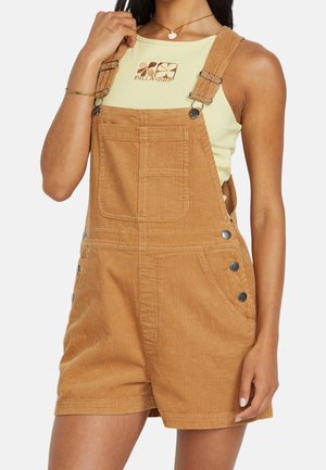 Kvinde iført lysegul ribbet tanktop med blomsterlogo og brune kordshorts med metalknapper og frontlomme.