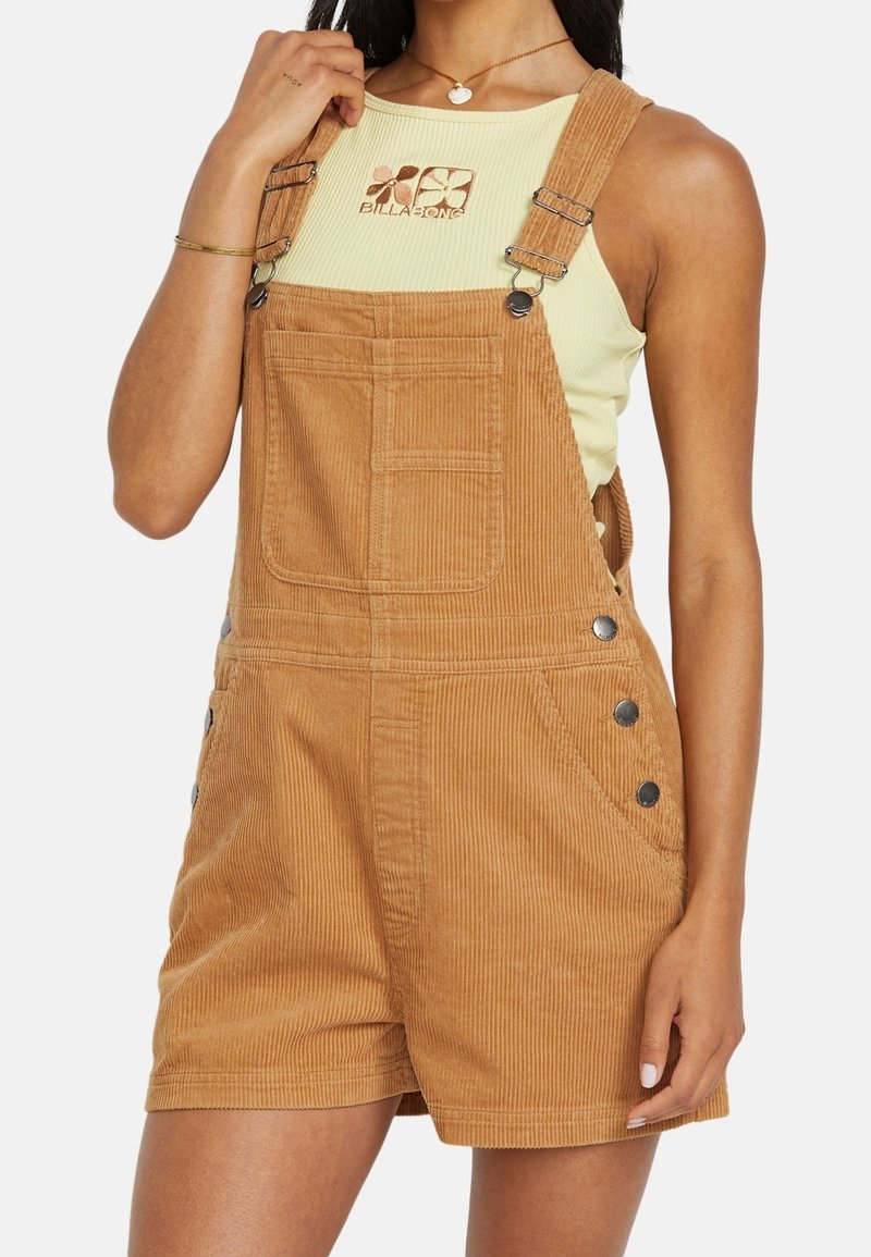 Femme portant un débardeur côtelé jaune clair avec un logo floral et une salopette courte en velours côtelé beige avec boutons métalliques et poche avant.