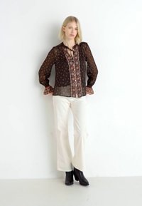 Blusa nera trasparente con motivo floreale, scollatura a chiave e polsini arricciati, abbinata a pantaloni bianchi a zampa d'elefante e stivaletti neri alla caviglia.