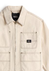 Veste beige Vans avec coutures contrastantes foncées, fermeture éclair à l'avant et trois poches plaquées à l'avant, dont une avec un patch logo Vans.