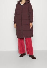 Abrigo tipo acolchado burdeos con mangas largas, capucha y cremallera frontal. Presenta costuras acolchadas horizontales y bolsillos laterales, usado sobre unos pantalones de pana rojos.
