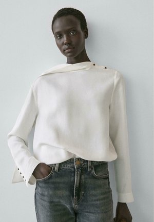 Dunkelhäutige Person trägt ein weißes asymmetrisch geknöpftes Oberteil und ausgewaschene blaue Jeans, steht vor einer schlichten hellgrauen Wand.