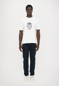 Camiseta blanca de algodón con un gráfico colorido de calavera en la parte frontal, combinada con pantalones oscuros de corte slim y zapatillas blancas.