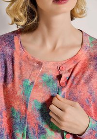 Frau, die den obersten Knopf einer bunten, strukturierten Strickjacke mit Farbnuancen von Rot, Grün und Lila schließt, wobei nur Hals und Kinn sichtbar sind.