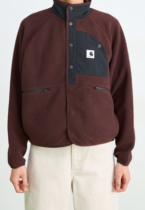 Brązowa kurtka polarowa z czarną patką na zatrzaski i kieszenią na piersi, noszona z jasnobeżowymi spodniami, ręce swobodnie wzdłuż ciała.