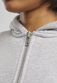 Gros plan d'une personne portant un sweat à capuche gris clair avec une fermeture éclair métallique argentée fermée jusqu'au cou.