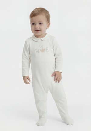 Småbarn med lyst brunt hår iført en hvit heldrakt med dyreillustrasjoner på brystet, stående mot en ensfarget bakgrunn.