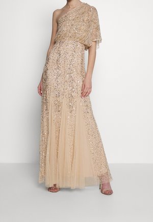 Beżowa, długia do ziemi suknia na jedno ramię, ozdobiona błyszczącymi cekinami i z płynącym tiulowym dołem, noszona z beżowymi sandałami na wysokim obcasie.