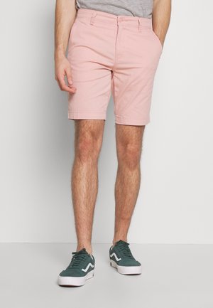 Mann trägt hellrosa Shorts, graues Shirt und dunkelgrüne Sneaker und steht vor einem einfarbigen hellen Hintergrund.