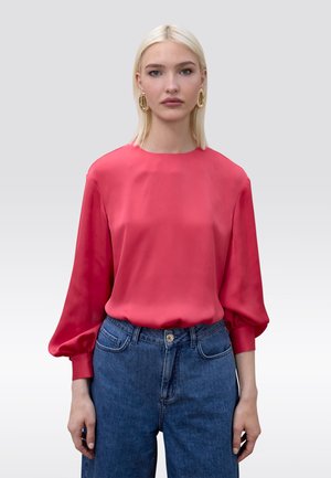 Mujer rubia con grandes pendientes ovalados dorados, una blusa rosa de manga larga con puños abullonados y jeans azules de cintura alta sobre un fondo gris.