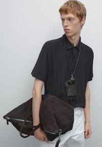 Černé polo tričko s ribovanou texturou, spárované s tmavou semišovou taškou na doklady, která má stříbrné zipy. Z náhrdelníku visí malý tmavý držák.