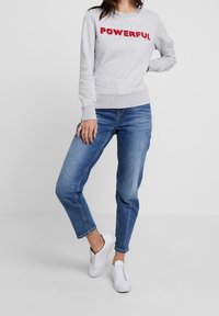 Sudadera gris clara con texto rojo "PODEROSO", combinada con jeans azules y zapatillas blancas. El corte es relajado con puños y dobladillo acanalados.
