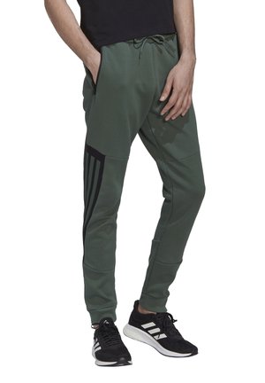 Man die groene taps toelopende joggerbroek draagt met zwarte zijstrepen en zwart-witte sportschoenen, staand met handen in de zakken.