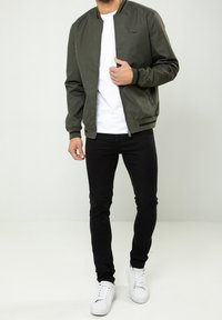 Homme portant une veste bomber vert olive par-dessus une chemise blanche, un jean skinny noir et des baskets blanches, debout devant un fond uni.