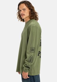 Man with curly hair wearing an olive green long-sleeve shirt with black text and logos on the sleeve and back, paired with blue jeans.