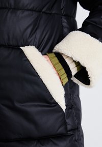 Čierna bundu typu puffer s krémovým umelým kožúškom, na ktorej je vidieť ruku oblečenú v zelenom pruhovanom svetri, ktorá vykúka z vrecka.