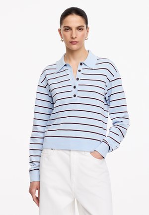Femme debout face à l'avant, portant un pull bleu clair à col et rayures foncées et un pantalon blanc, une main dans la poche.