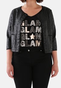 A woman wears a black, patterned jacket over a glittering "GLAM" graphic t-shirt and black pants, posing with her hands by her sides.