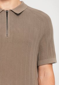 Nærbilde av en person som har på seg en kortermet, ribbestrikket polo-skjorte i taupe med glidelås foran.