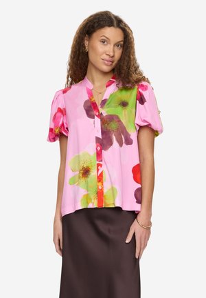 Kobieta o kręconych włosach, ubrana w różową, kwiatową bluzkę z krótkim rękawem i ciemnobrązową spódnicę, stojąca na białym tle.