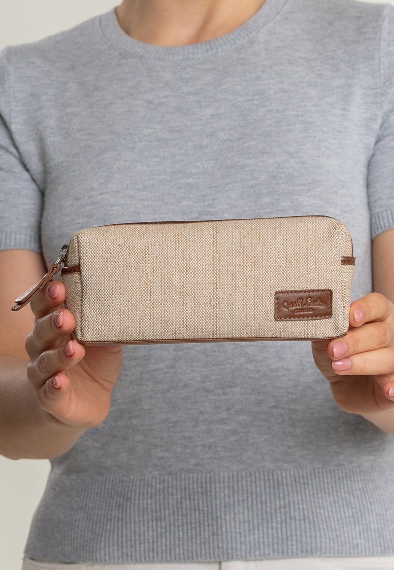 Beige tyg penna fall med en texturerad yta, bruna läderdetaljer och dragkedjestängning. Har en varumärkeslogotyp på sidan.