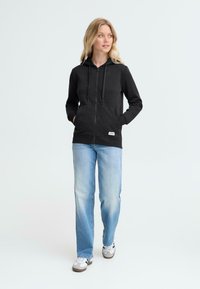 Svart hettegenser med glidelås, frontlommer og snøring, sammen med lyseblå jeans og sneakers, foran en ensfarget bakgrunn.
