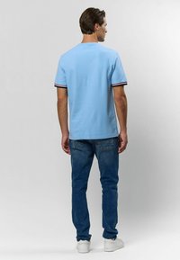 Svetlomodrá košeľa s krátkym rukávom a kontrastnými čiernymi a červenými pruhmi na manžetách, skombinovaná so sivomodrými džínami a bielymi teniskami, zachytená zozadu.