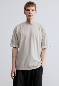 Beiges T-Shirt mit kurzen Ärmeln aus weichem Stoff, mit rundem Ausschnitt und gestreiften Akzenten an den Bündchen. Getragen zu lockeren schwarzen Hosen.