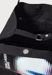 Czarna torba na ramię z tkaniny płóciennej, wyposażona w górne zapięcie na zatrzask, wewnętrzną kieszeń na zamek oraz przedni nadruk graficzny z żywymi akcentami kolorystycznymi.