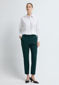 Witte cropped blouse met lange mouwen en een fitted ontwerp, gecombineerd met donkergroene pantalon en zwarte hoge hakken.