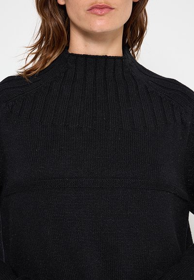 Femme portant un pull en maille côtelée noir à col montant, montrant le haut du torse et une partie de son visage.