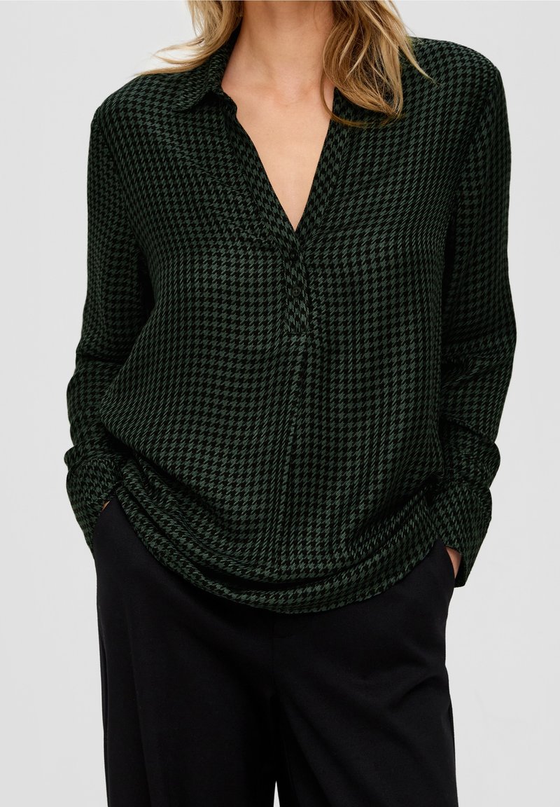 Femme portant un chemisier à manches longues pied-de-poule vert foncé et noir avec col en V et pantalon noir, mains dans les poches, sur fond uni.