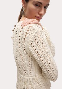 Sweter z dzianiny w kolorze kremowym z teksturowanym wzorem, z otwartymi detalami. Projekt zawiera frędzle na dole.