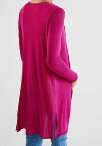 Fuchsia långärmad tunika med lös passform, sidofickor och rundad halsringning, kombinerad med blå jeans. Mjuk, lätt tygtextur.